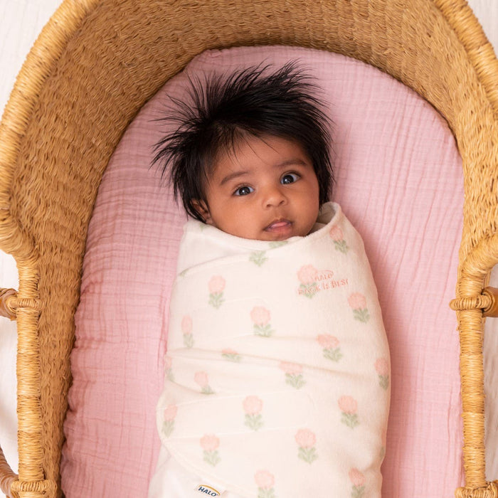 A dark-haired baby is snugly wrapped in a HALO Swaddle Micro-Fleece TOG 3.0 with adjustable wings, featuring a floral pattern. Nestled in a pink-lined wicker basket, the babys wide eyes gaze upward.