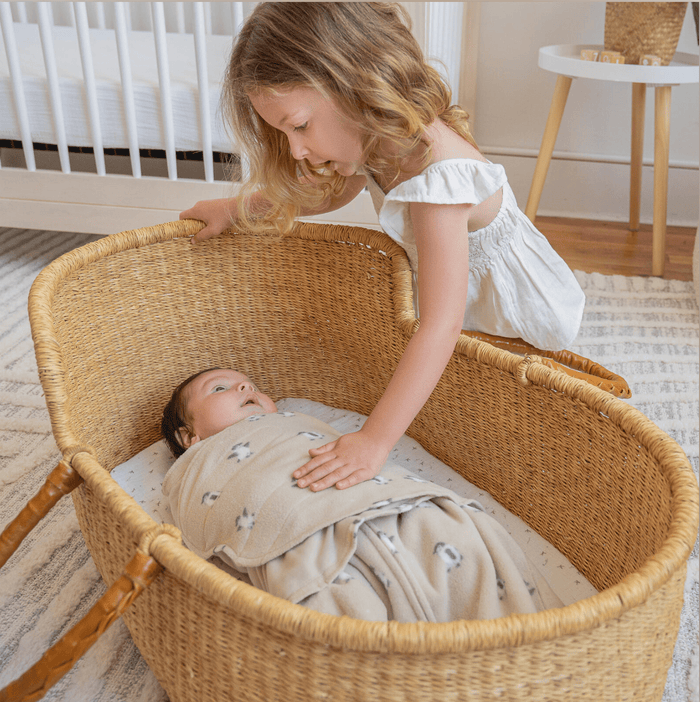 A young girl with curly hair, in a white dress, gently places her hand on a baby wrapped in a HALO Swaddle Micro-Fleece TOG 3.0, lying in a wicker bassinet. The room features a crib, small table, and patterned rug—ideal for healthy hip development with its adjustable wings.