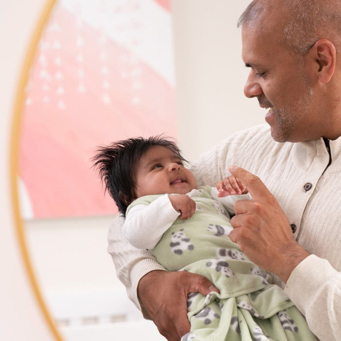 A person smiles while holding a baby in a green HALO Swaddle Micro-Fleece TOG 3.0, featuring paw prints. The content baby, enjoying adjustable wings for healthy hip development, gazes up at the person. Blurred pink and white artwork is visible in the background.