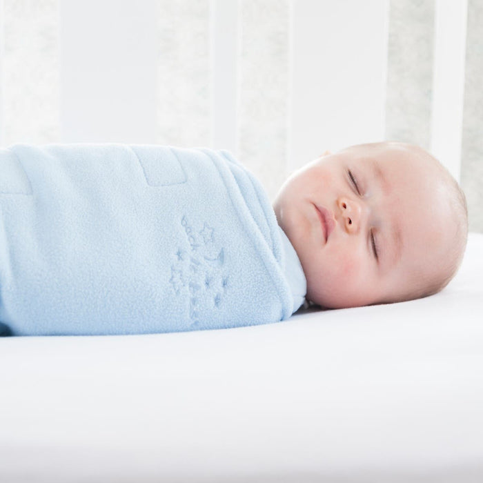 A baby sleeps serenely in a HALO Swaddle Micro-Fleece TOG 3.0, wrapped in a starry light blue blanket. Resting on a white mattress and surrounded by crib rails, adjustable wings provide comfort and support for healthy hip development as soft light filters in.