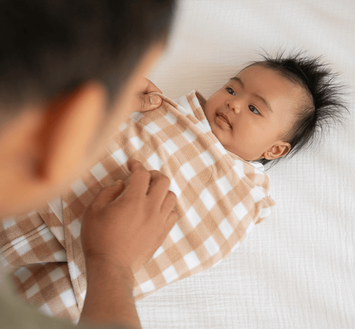 A parent swaddles a baby in HALOs Swaddle Micro-Fleece TOG 3.0, renowned for promoting healthy hip development. The cozy, checkered wrap features beige and white squares, keeping the baby content and relaxed on a white surface.