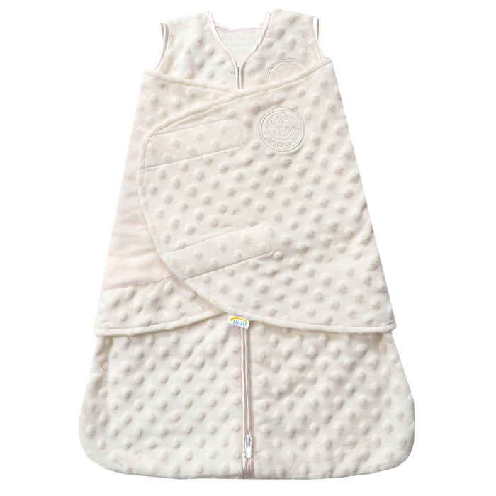 The HALO Swaddle Plushy Dot Velboa TOG 3.0 is a cream-colored, sleeveless SleepSack® with a soft, dot pattern. It features a bottom zip closure, adjustable wings for healthy hip development, and an embroidered logo on the wrap-around flap.