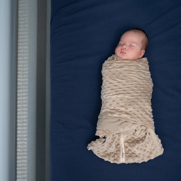 A newborn sleeps peacefully, wrapped in a HALO Swaddle Plushy Dot Velboa TOG 3.0, and rests on a textured beige blanket over a dark blue sheet.