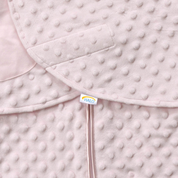 A close-up of the Swaddle Plushy Dot Velboa TOG 3.0 baby blanket shows its soft, textured pink fabric with small circular bumps. It features a HALO tagged zipper pull and adjustable wings, designed for healthy hip development as part of the HALO SleepSack® Swaddle collection.