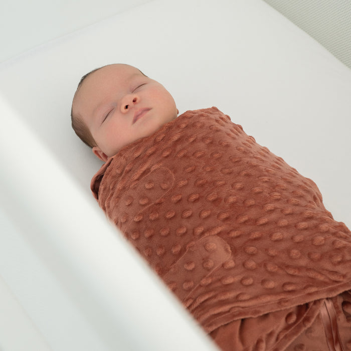 A sleeping baby is swaddled in a brown HALO Swaddle Plushy Dot Velboa TOG 3.0 and lies on a white mattress inside a crib.