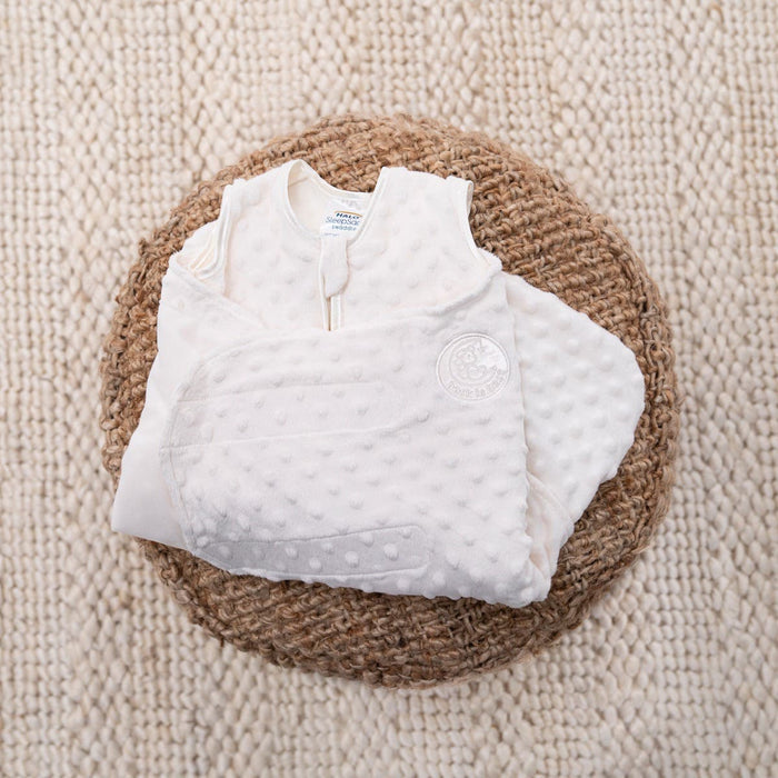 The HALO Swaddle Plushy Dot Velboa TOG 3.0, designed for healthy hip development, is softly textured and elegantly folded on a round woven rug against a cream-colored knitted backdrop.