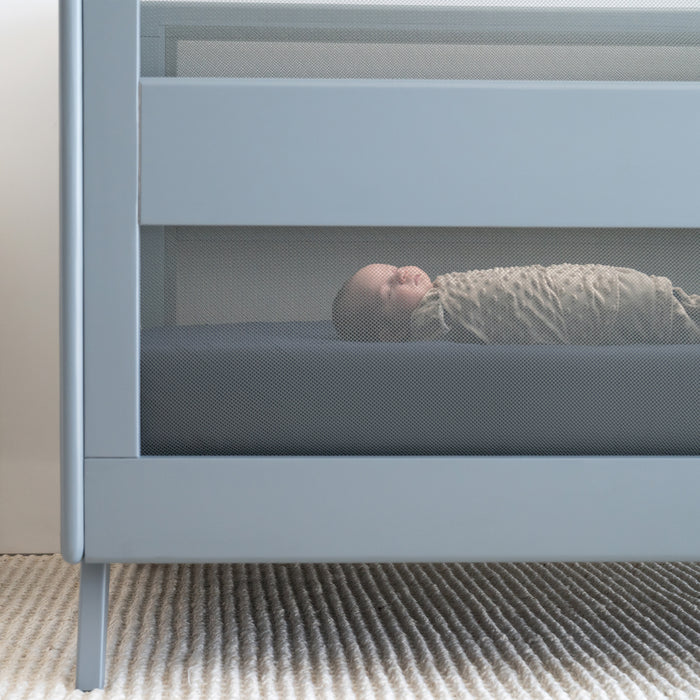 A newborn wrapped in the HALO Swaddle Plushy Dot Velboa TOG 3.0 rests on their back in a modern gray crib with mesh sides, set atop a beige textured rug.