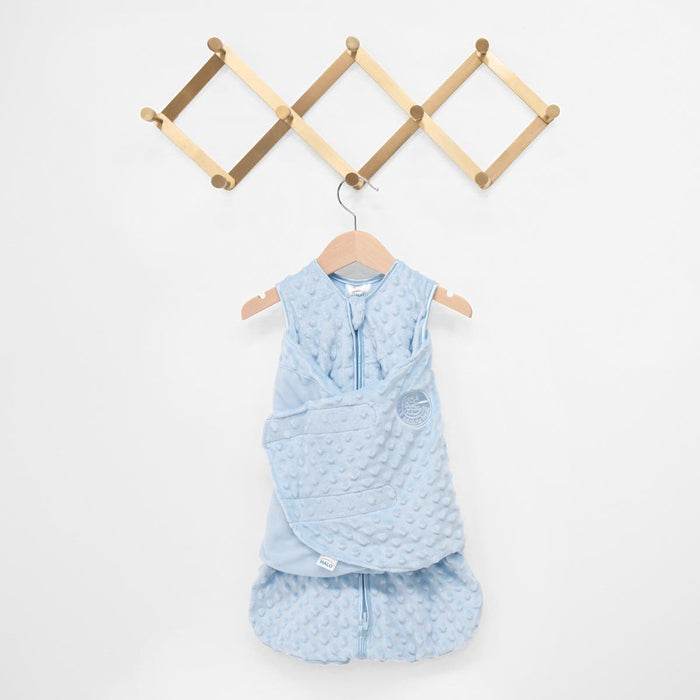 A HALO Swaddle Plushy Dot Velboa TOG 3.0 in light blue with a zip front, soft dotted texture, and adjustable arm wrap for healthy hip development is hanging on a wooden coat rack against a white wall.