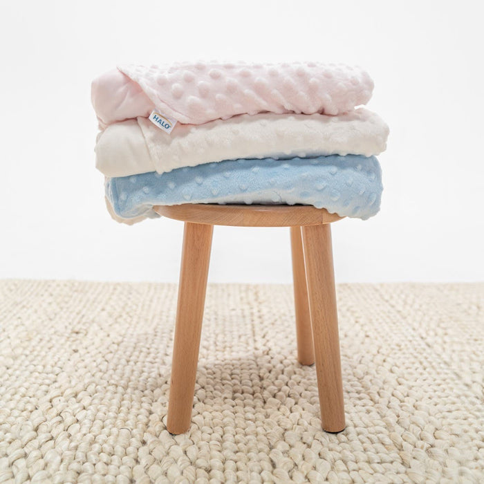 Three soft Velboa TOG 3.0 baby blankets in pink, white, and blue are folded on a small wooden stool with a HALO SleepSack® Swaddle nearby. The stool rests on a woven beige rug against a plain white backdrop.