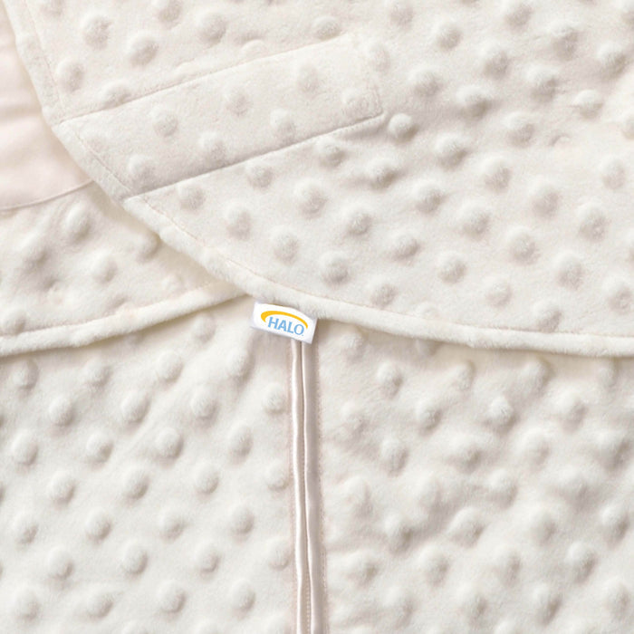 A close-up of the textured, cream Swaddle Plushy Dot Velboa TOG 3.0 fabric by HALO reveals raised dots and a small HALO tag at the overlapping seam with adjustable wings for healthy hip development.