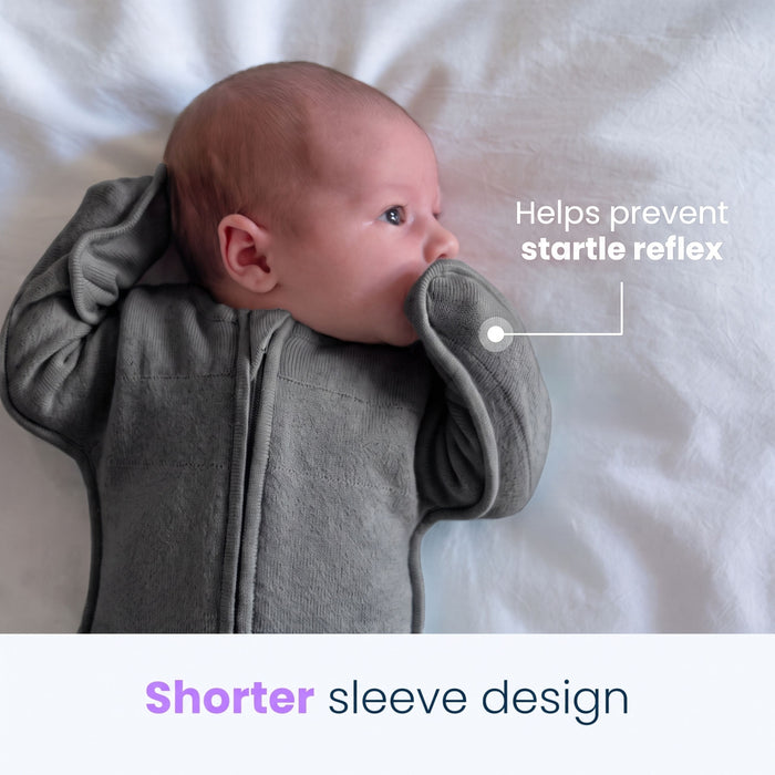 A baby wearing the HALO SwaddleStretch™ Newborn Swaddle TOG 0.5 with shorter sleeves rests on a white blanket. Text reads: Helps prevent startle reflex and Shorter sleeve design. The infant’s hands are positioned near their face, highlighting the hip-healthy sleep sack, designed for comfortable and secure swaddling.