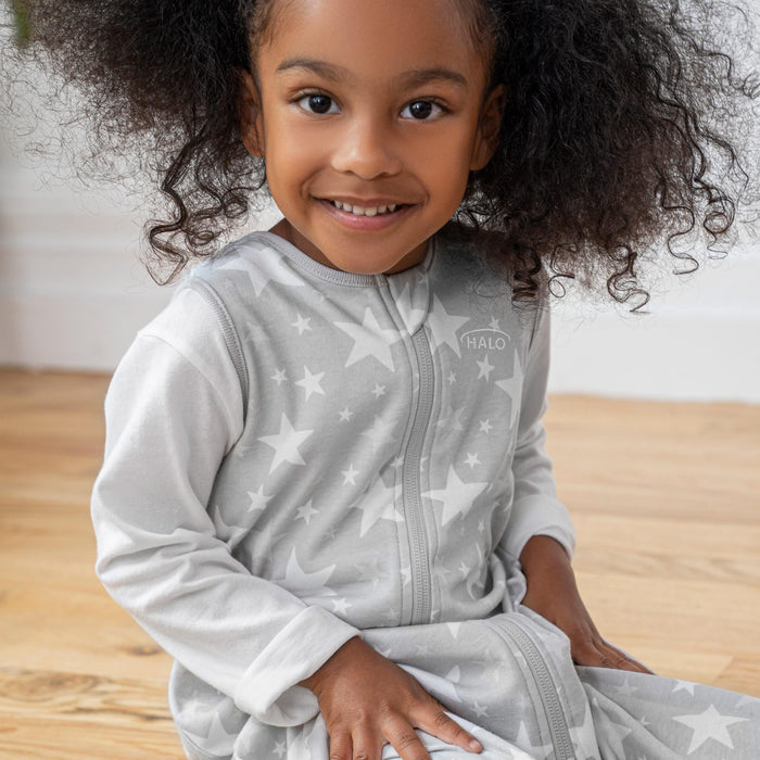 A curly-haired toddler smiles while sitting on the wooden floor, wearing a HALO Toddler 100% Cotton TOG 0.5 SleepSack, light gray with white stars and long sleeves, featuring a zipper. A simple white wall in the background adds to the cozy ambiance.