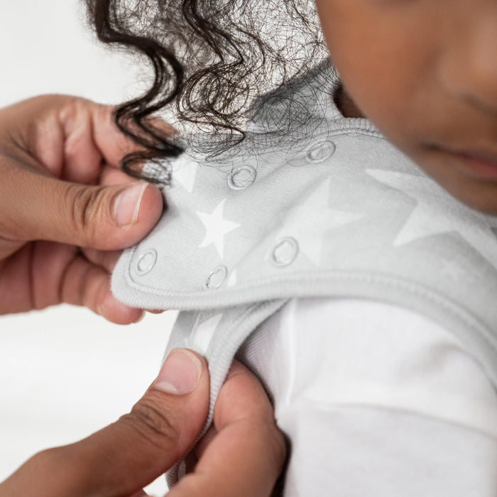 A person adjusts the shoulder snaps on a toddlers HALO SleepSack, featuring a light gray 100% cotton fabric with a white star pattern. The curly-haired child in a white shirt sits patiently while the focus remains on the hands and adjustable features of this cozy TOG 0.5 garment.