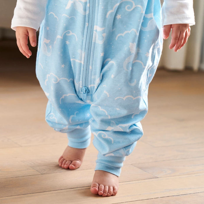 A toddler on a wooden floor wears a blue onesie with cloud and moon patterns, echoing their HALO Toddler 100% Cotton TOG 0.5. Barefoot and ready for bedtime, the scene is bright and cozy with the onesies adjustable features ensuring comfort as they drift into sleep.