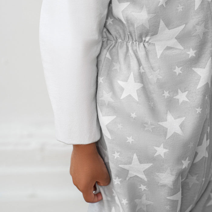 A close-up shows a toddler in a HALO Toddler 100% Cotton TOG 0.5 sleepsack with a white star pattern on gray. Only the arm and torso are visible, highlighting the soft, cozy fabric, set against a plain light background.