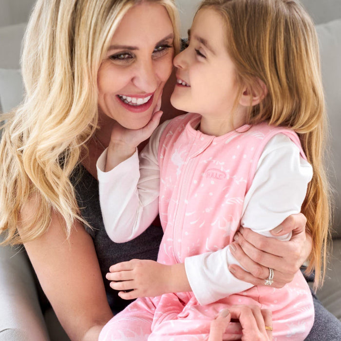 A smiling woman with long blonde hair holds a young girl in her lap, who gently touches the womans face. The girl, dressed in a pink and white outfit, sits comfortably on the light-colored couch wrapped snugly in her HALO Toddler 100% Cotton TOG 0.5 for better sleep with adjustable features.