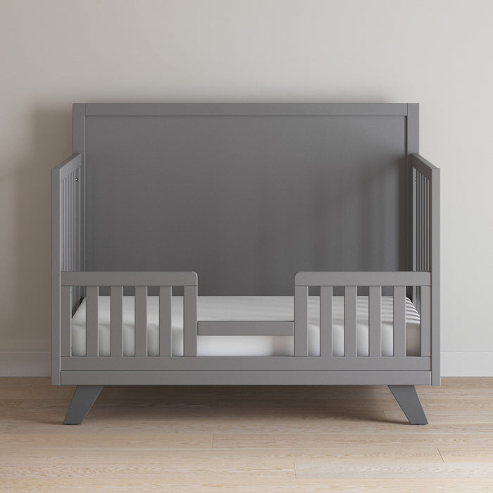 The Child Craft Toddler Guard Rail in Cool Gray is shown against a plain light wall on a light wood floor, featuring open sides, a low front guardrail, and a white mattress with a striped bedsheet.