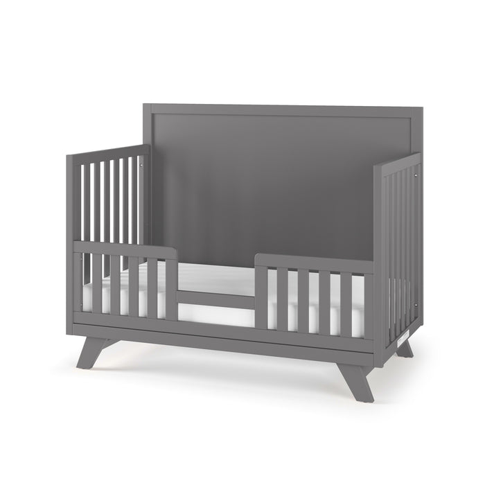 The Child Craft Toddler Guard Rail in Cool Gray features a high solid headboard, open slatted sides, low guardrails on each side, a white mattress, and sits on angled legs.