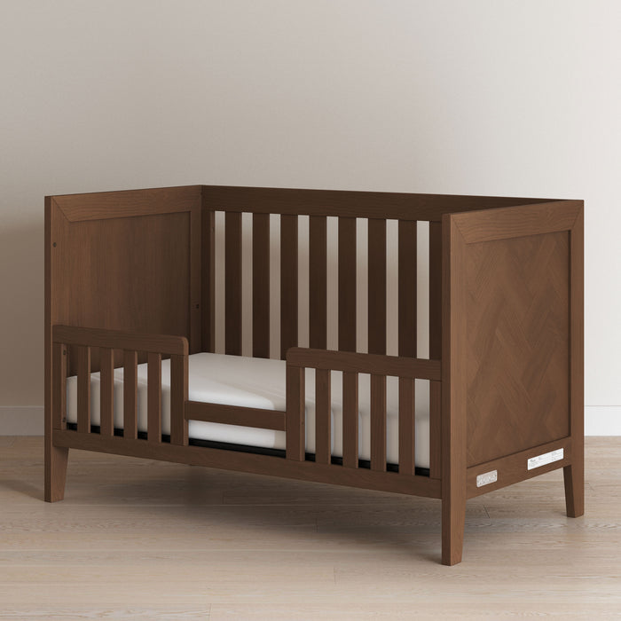 A Child Craft Toddler Guard Rail in Toasted Chestnut is installed on a wooden toddler bed, featuring a baby-safe finish and a white mattress, placed on a light wood floor against a plain, light-colored wall.