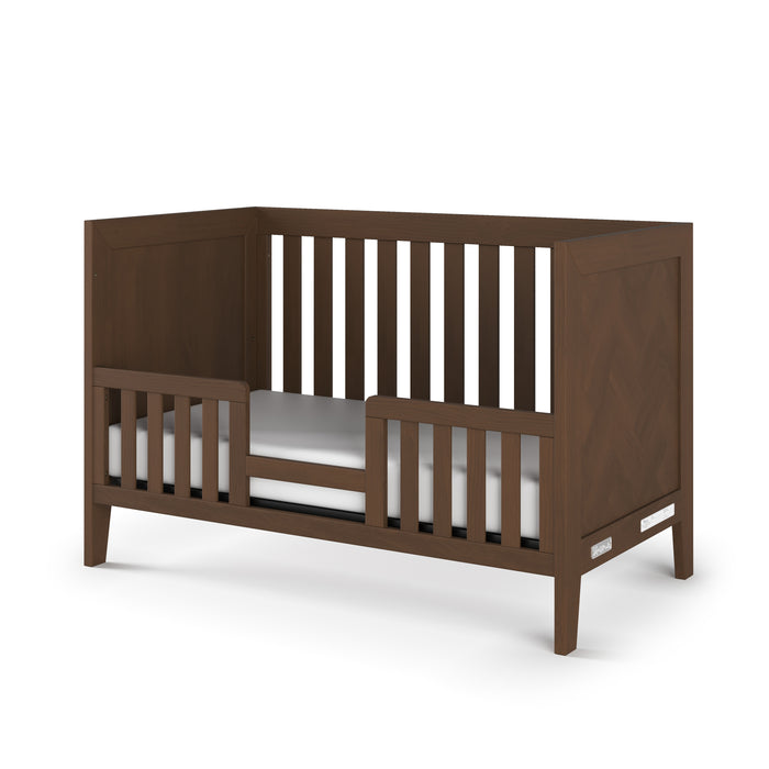 The Child Craft Toddler Guard Rail in Toasted Chestnut features baby-safe finishes, a modern minimalist design with open side and back slats, a low front guard rail, and includes a white mattress.