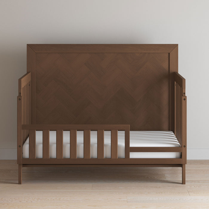 The Child Craft Toddler Guard Rail in Toasted Chestnut features slatted sides, a high textured headboard, and a partial guard rail. Finished baby-safe, it stands on a light wood floor against a white wall and holds an uncovered white mattress.