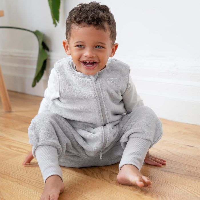 A toddler in a light gray, sleeveless onesie with a zipper sits playfully on a wooden floor, mirroring the comfort and style of HALOs Toddler Luxe Fleece TOG 2.0. Smiling with curly hair, he is framed by a white wall and green plant that enhance the calm, safe ambiance.