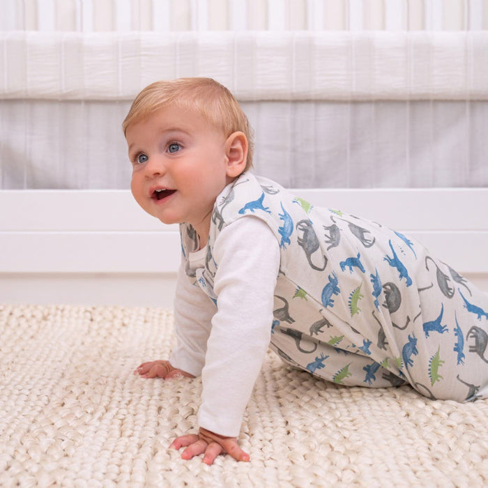A baby with blonde hair crawls on a cream woven rug in a white outfit featuring blue, green, and gray dinosaurs. A white crib and wall provide a safe sleep setting embraced by cozy 100% cotton textures from HALOs Wearable Blanket TOG 0.5.