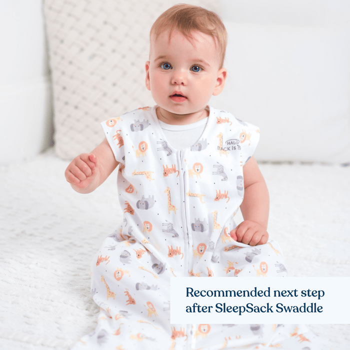 A baby sits on a white blanket in a HALO Wearable Blanket 100% Cotton TOG 0.5 adorned with giraffes and lions, perfect after the SleepSack Swaddle. A neutral-toned pillow in the background emphasizes safe sleep with this breathable sleep sack.
