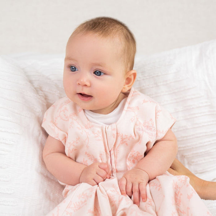 A baby with light hair and blue eyes sits on a white textured blanket, wearing a light pink patterned outfit. Their curious gaze drifts sideward while they enjoy their HALO Wearable Blanket 100% Cotton TOG 0.5, ensuring safe sleep with its cozy design.