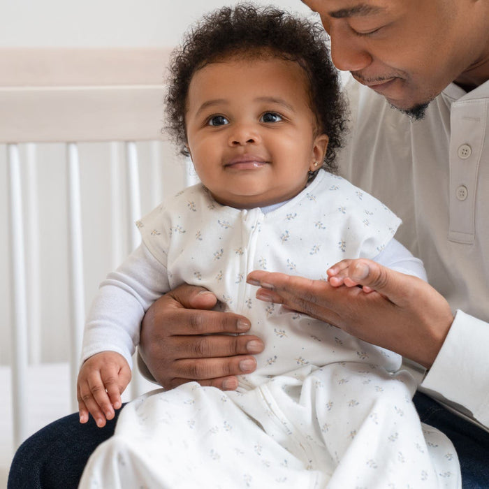 A smiling baby, in a white patterned outfit, sits on an adults lap in a cozy room. The adult, wearing a white shirt, gently holds and looks at the baby beside a white crib, ensuring safe sleep with HALOs Wearable Blanket 100% Cotton TOG 0.5.