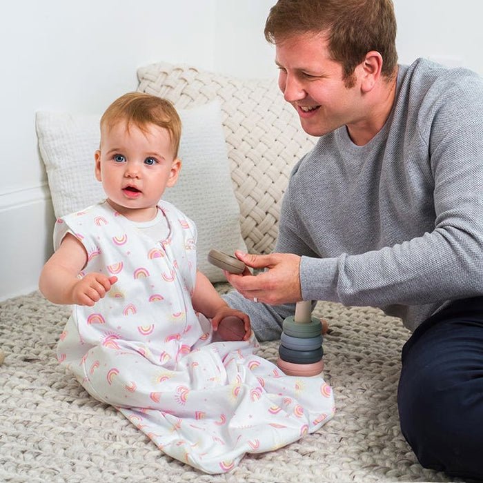 A smiling adult sits on a light gray rug, holding toy rings, while looking at a baby in HALOs Wearable Blanket 100% Cotton TOG 0.5 with rainbow patterns. The baby is seated on the rug, surrounded by textured white pillows, enjoying safe sleep and playtime.