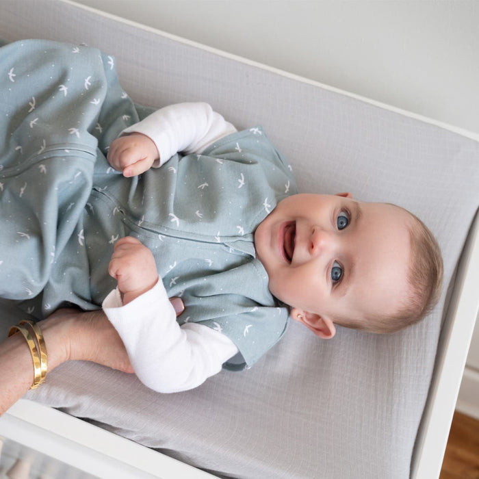 A baby in a light blue outfit with small white bird patterns lies on a changing table wrapped in the HALO Wearable Blanket 100% Cotton TOG 0.5. The baby smiles while looking up, supported by a gentle hand to ensure safe sleep.