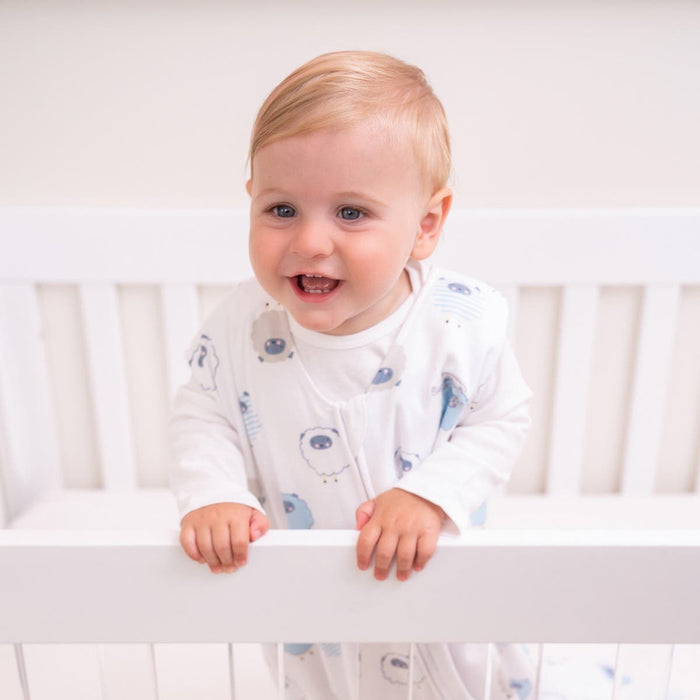 A joyful baby with light hair, wearing a sheep-patterned white onesie and wrapped in HALOs Wearable Blanket 100% Cotton TOG 0.5, stands cheerfully in the crib, ensuring playful moments and safe sleep.