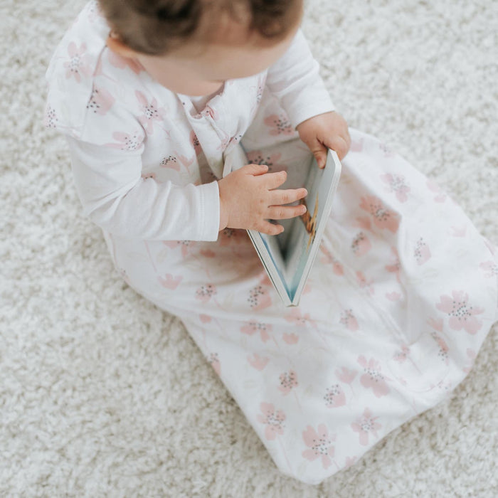 A baby in a HALO Wearable Blanket, made of 100% cotton with pink floral patterns, sits on a soft, light-colored carpet, gazing at and touching a book.