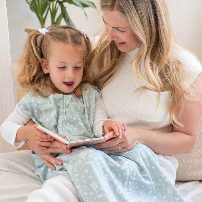 In a cozy, well-lit space, a woman in a white top and a young girl with pigtails in a light blue patterned dress are engrossed in reading. Nearby, the comfort of the HALO Wearable Blanket 100% Cotton TOG 0.5 ensures safe sleep for little ones later on.