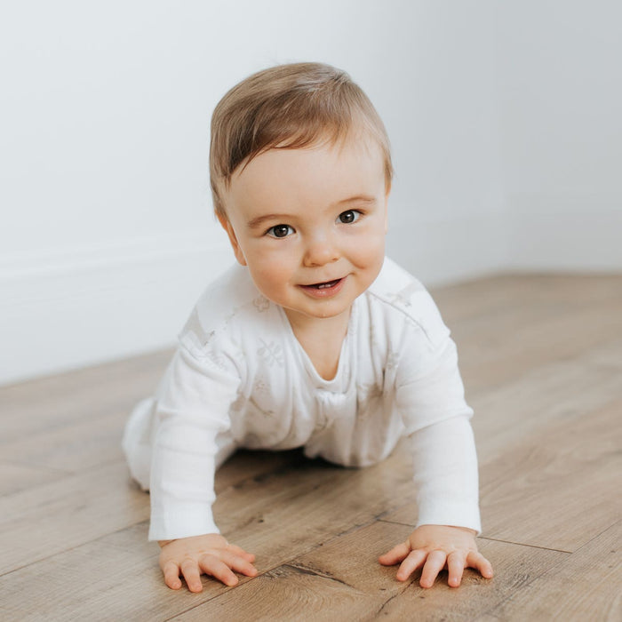 A baby in a white onesie crawls on a wooden floor, smiling at the camera. Nearby lies a HALO Wearable Blanket 100% Cotton TOG 0.5, ready for safe sleep against a plain white wall.