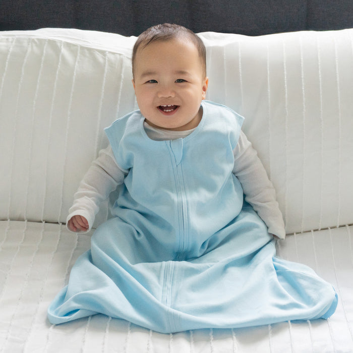 A smiling baby in a HALO Wearable Blanket 100% Organic Cotton TOG 0.5 sits on a white, quilted bed with a gray headboard, highlighting comfort and safe sleep.