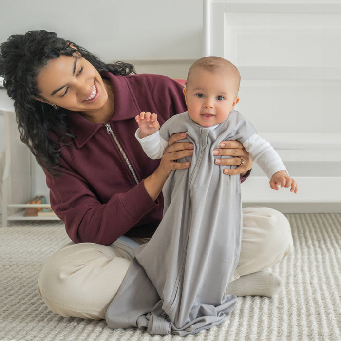 A smiling adult holds a happy baby sitting upright. The baby wears the HALO Wearable Blanket 100% Organic Cotton TOG 0.5 over a white shirt, in a softly lit, cozy nursery with neutral tones.