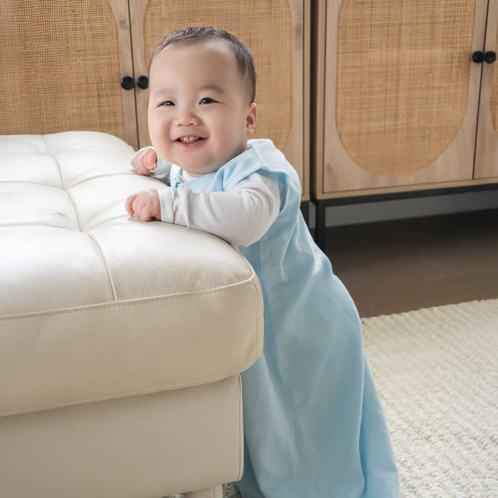 A smiling baby stands by a white ottoman wearing the HALO Wearable Blanket 100% Organic Cotton TOG 0.5 over a long-sleeve shirt. The cozy room includes a cabinet with woven panels and a cream rug, creating a comfortable, safe sleep space.