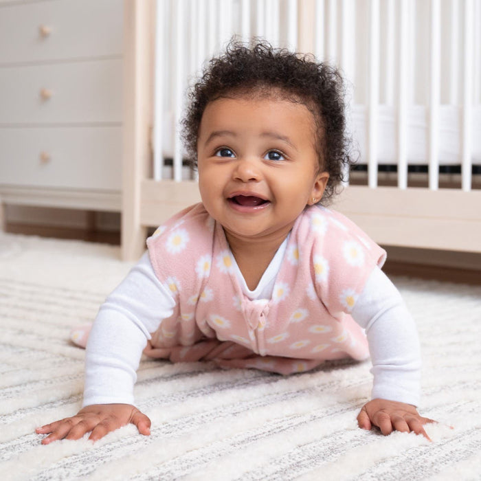 A happy baby with curly hair crawls on a light rug in a nursery, wrapped in a pink HALO Wearable Blanket Micro-Fleece TOG 1.0 with white daisies, promoting safe sleep as a crib and dresser grace the background.