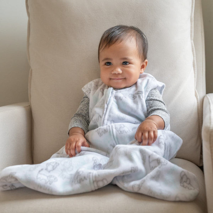 A baby with dark hair smiles while sitting on a beige armchair, wrapped in HALOs cozy Wearable Blanket Micro-Fleece TOG 1.0 in a gray and white pattern, creating a warm, inviting atmosphere with its softly lit chair and background.