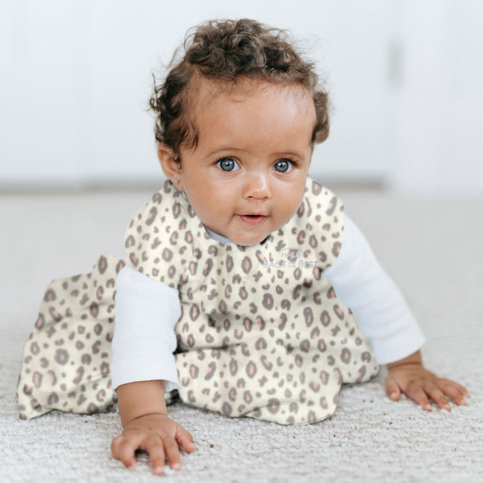 A curly-haired baby with bright eyes, wearing a leopard print outfit and a HALO Wearable Blanket Micro-Fleece TOG 1.0, sits on a carpeted floor, curiously looking at the camera. The softly blurred background enhances the gentle ambiance.