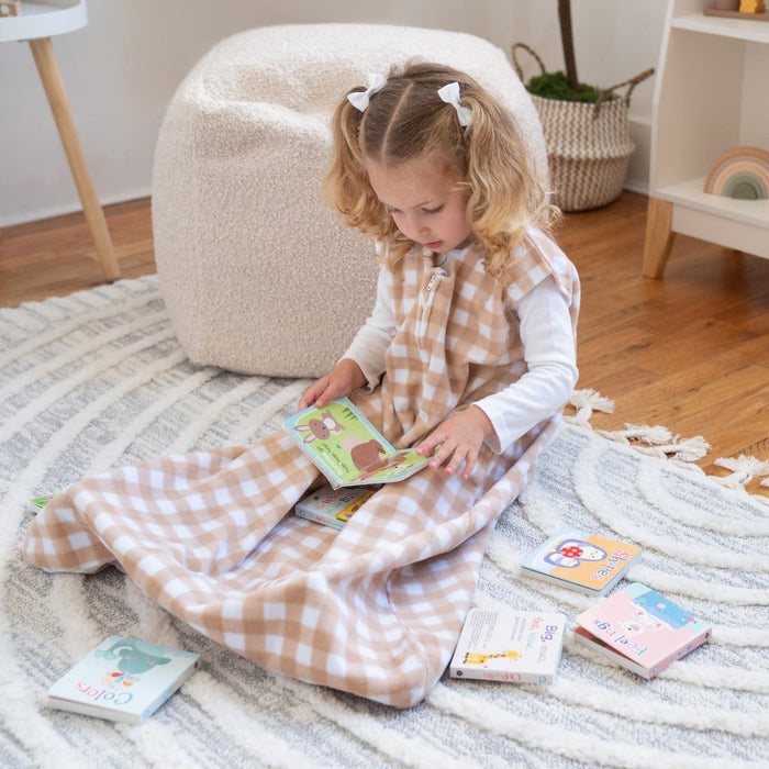 A young child with curly hair sits on a circular rug, wrapped in a HALO Wearable Blanket Micro-Fleece TOG 1.0. They are reading a book, surrounded by other childrens books. Bedroom furniture and decor create a warm backdrop for comfort and safe sleep.