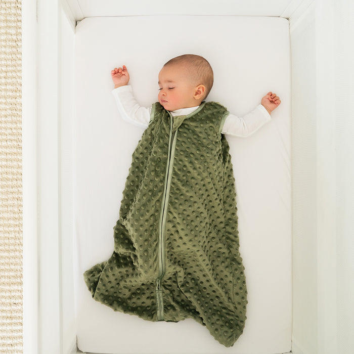 A baby sleeps peacefully on their back in a white crib, wearing the HALO Wearable Blanket Plushy Dot Velboa TOG 1.5 in soft green, with outstretched arms and a front zipper for comfort.